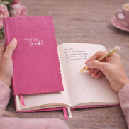 Donna che scrive su un taccuino A5 rosa “Think Pink!” su una scrivania, scena lifestyle elegante e naturale per lavoro e creatività