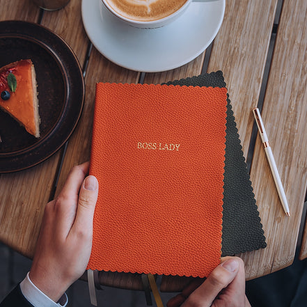 Taccuino Boss Lady formato A5 con copertina vegana arancione utilizzato su una scrivania con tazza di caffè e penna, ideale per lavoro creativo e momenti di ispirazione
