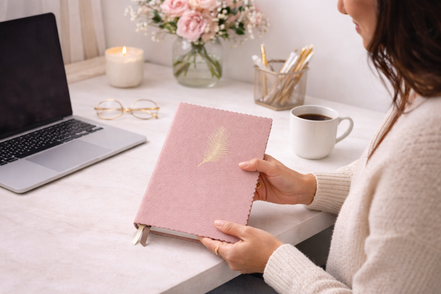 Donna seduta a una scrivania bianca e ordinata mentre utilizza un taccuino rosa elegante, in un ambiente luminoso e minimal, stile lifestyle.